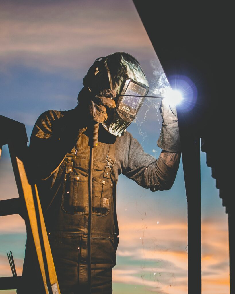 A person wearing a protective welding helmet and coveralls is welding a metal structure. They are holding a welding torch, with visible sparks and a bright light at the point of contact. A ladder is partially visible on the left side, and the sky is colorful at dusk, highlighting the beauty of engineering & design in action.