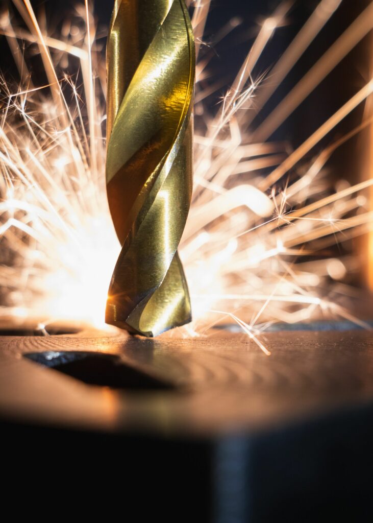 A close-up of a golden drill bit cutting through metal, generating bright, orange sparks that scatter in various directions. The intricacies of fabrication are evident as the drill bit and the sparks remain sharply in focus, while the background is dark and blurred, emphasizing the action.