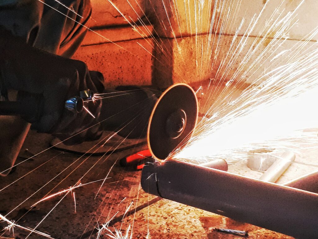 In a dimly lit workshop, a person uses an angle grinder to cut through a metal pipe, creating a shower of bright sparks. The background includes concrete surfaces and scattered tools, emphasizing the industrial setting typical of construction and installation environments.