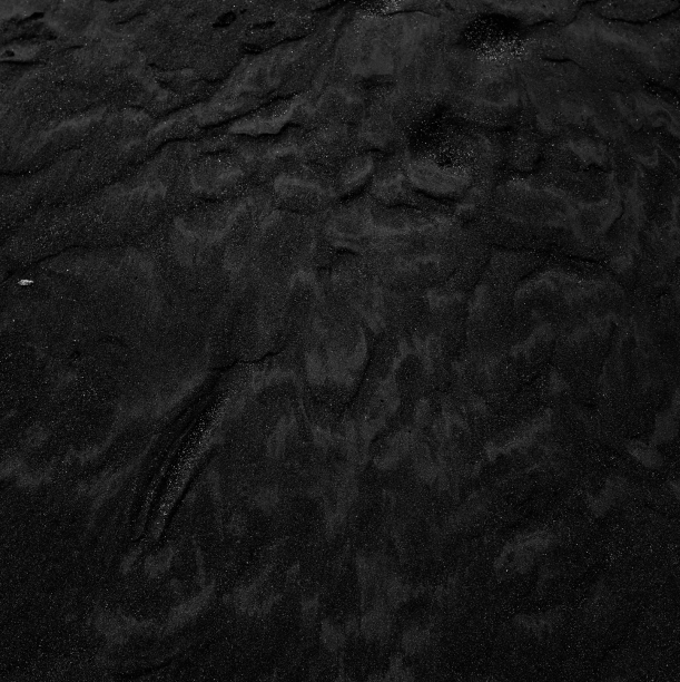 A close-up of dark, textured sand with various patterns and ridges lies undisturbed, its rough surface showcasing subtle variations in tone and shadow that highlight the natural formations—a testament to the intricate craftsmanship evident about MJK Industries.