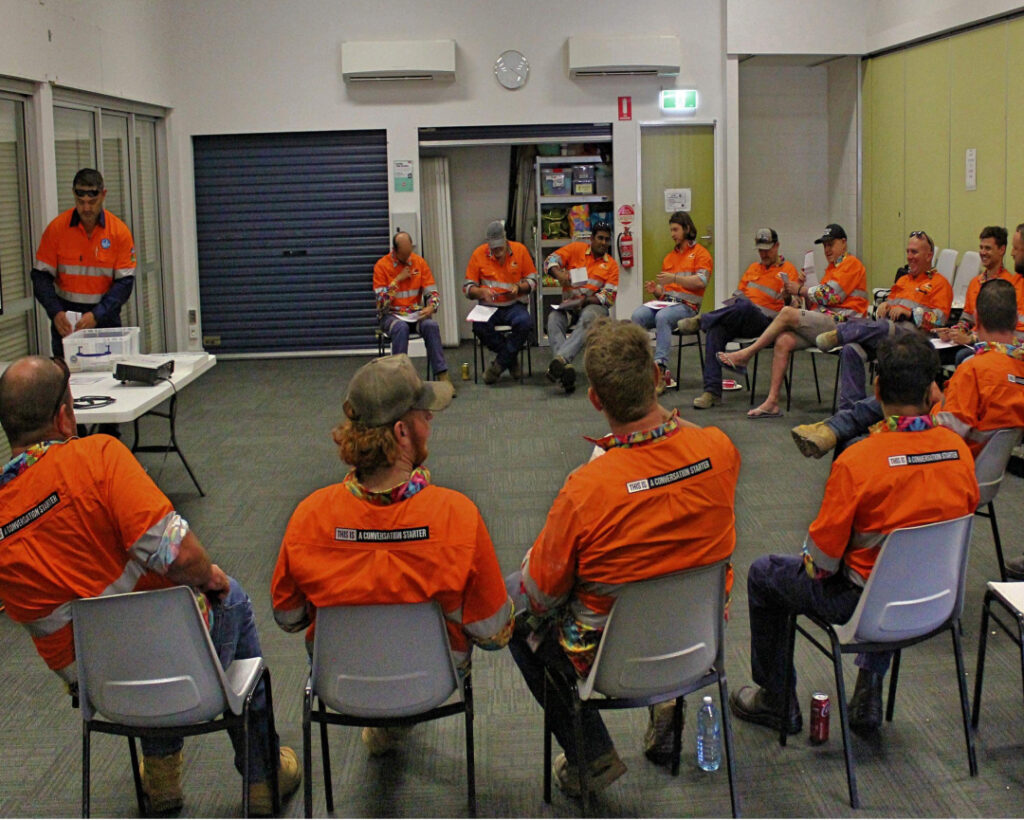 A group of workers in fluorescent orange shirts, likely SMP partners, sit in a circle in a room. They appear to be having a meeting. Some attendees are taking notes, while others are attentively listening. Safety gear and various personal items are visible. The room has air conditioning units on the walls.