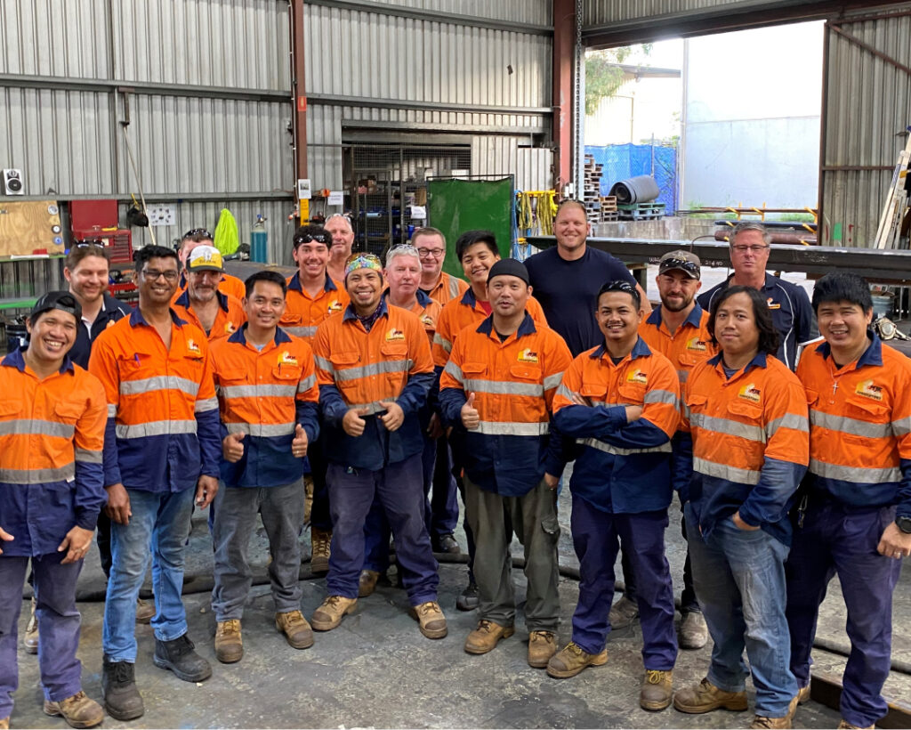 A group of 17 people, mostly men, are standing in an industrial workshop. They are wearing high-visibility orange and navy uniforms. Some are smiling for the photo. The workshop, a proud SMP partner, features metal beams, tools, and machinery in the background.