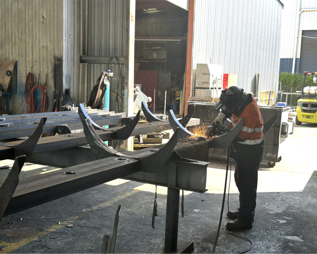 A worker, wearing protective gear including a helmet, collaborates with an SMP partner while using a welding torch, producing sparks. They are in an industrial outdoor setting filled with metal structures and equipment. Sunlight filters into the area, casting shadows on the ground.