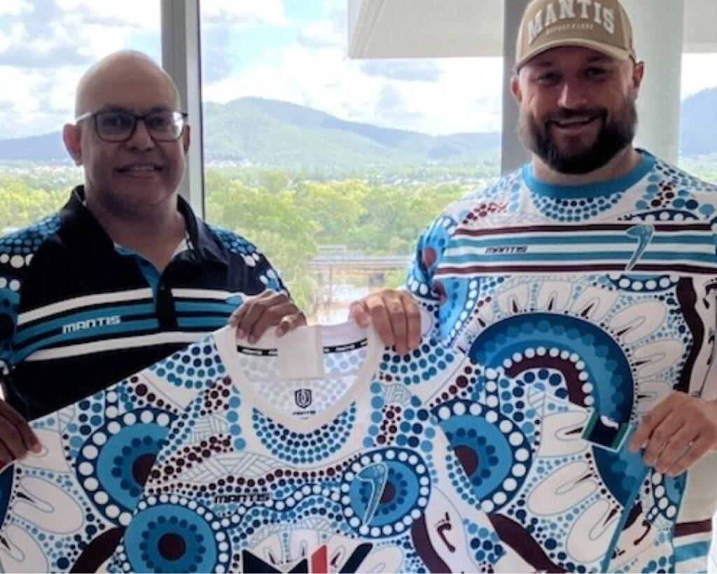 Two men are holding up and displaying Indigenous-designed rugby jerseys. The man on the left, an SMP partner, is wearing glasses and a black and blue polo shirt, while the man on the right is wearing a cap and a similar design shirt. Trees and mountains are visible in the background.