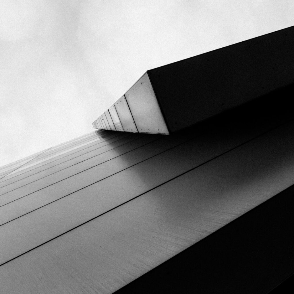 A black-and-white image of a modern, angular building taken from a low angle, accentuating its sharp lines and sleek geometric design. The perspective emphasizes the building's height against a cloudy sky background.
