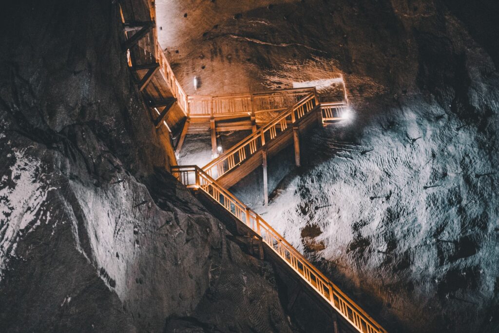 A wooden staircase with railings ascends through a dimly lit, rocky cave. The cavernous walls, reminiscent of mining and resources exploration, are rugged and textured, with light and shadow creating a dramatic ambiance. A few lights illuminate the path, adding a warm glow to the otherwise dark environment.
