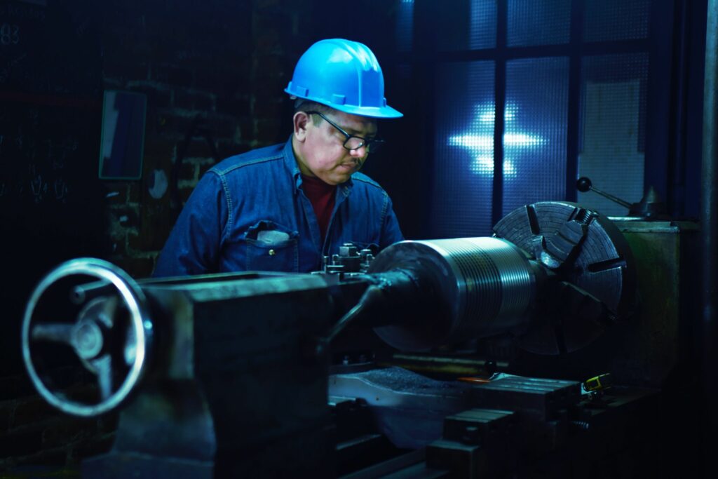 A worker wearing a blue hard hat and safety glasses operates a lathe machine in a dimly lit industrial setting. Focused on the task, the worker is contributing to critical infrastructure development, with a bright light visible in the background illuminating their precision.