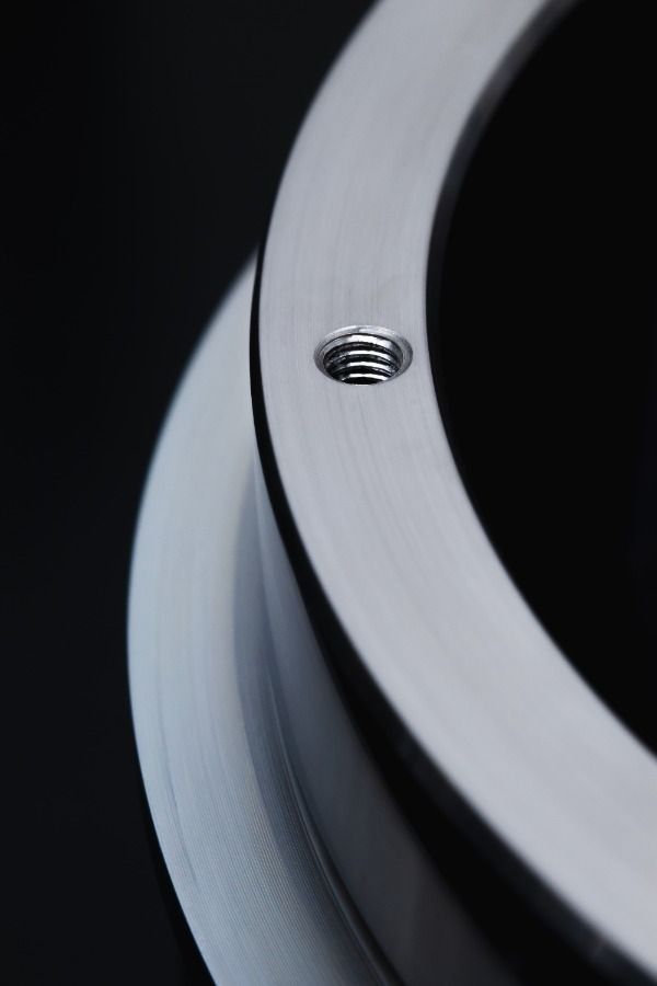 Close-up of a smooth, silver metallic circular object with a threaded hole on the surface, set against a dark background. The object appears to be a machined metal part, possibly fabricated from steel as a component of an industrial or mechanical assembly.