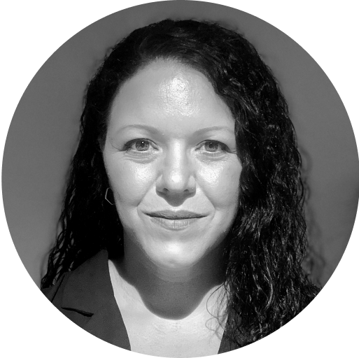 Black and white headshot of a person with long, curly dark hair, wearing a dark-colored top and light makeup, looking directly at the camera with a neutral expression. The image is circularly cropped, with a plain dark background.