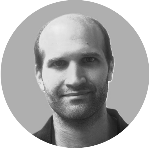 A portrait of a man with a slight smile, short dark hair, and a beard. He is wearing a dark-colored shirt, and the background is a plain light grey. The image has a circular frame.