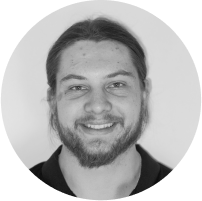 A black-and-white portrait photo of a person with long hair tied back. They are smiling and have a beard and mustache. They are wearing a dark-colored shirt and are set against a plain, light-colored background.