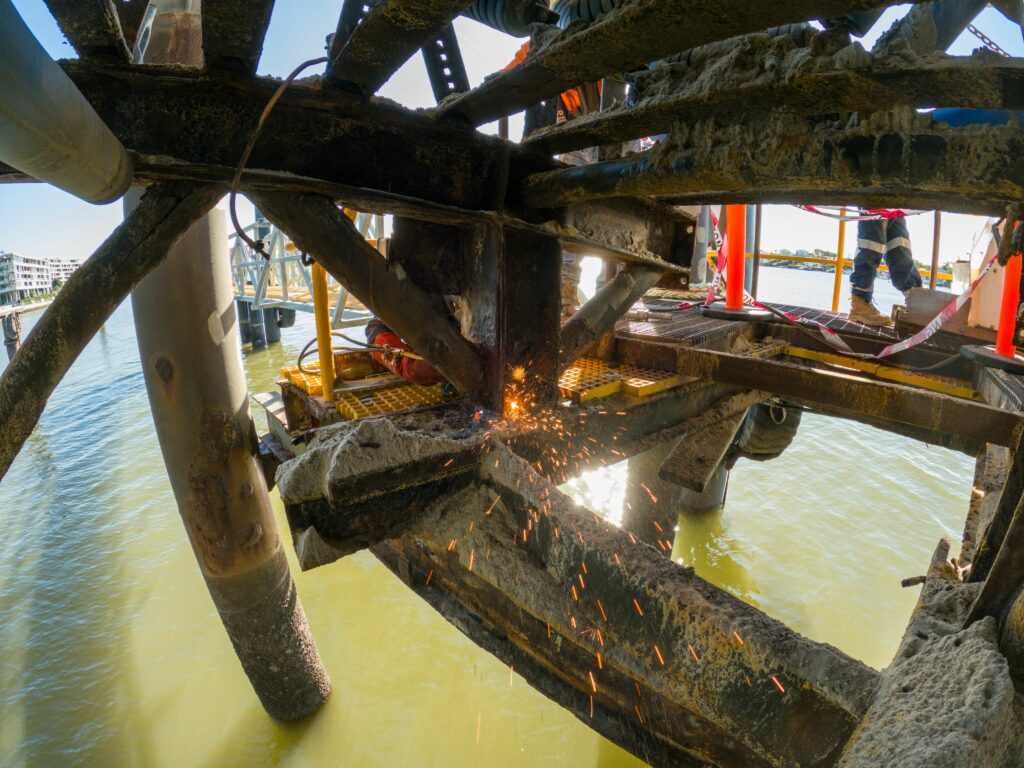 A construction worker is using a tool to fabricate and cut metal beams on a bridge extending over the water. Sparks are flying from the steel design. The worker is wearing protective gear, including a helmet and safety harness. The water below is calm and light green.