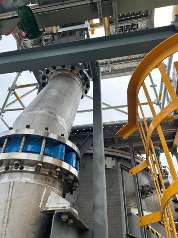 An industrial setup with large steel pipes and bolts extended towards a tall platform structure, seamlessly integrating recycling practices. Yellow stair railing is visible to the right, indicating access to elevated levels within this circular economy framework. The background shows parts of the metal framework and a cloudy sky.