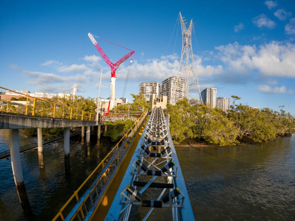 A bridge under construction extends over a body of water towards a cityscape with tall buildings. A large, red construction crane involved in materials handling and a power line tower are visible in the background, and there are trees along the water's edge under a partly cloudy sky.