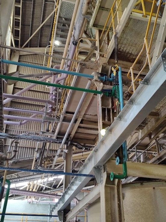 An industrial facility with a complex network of pipes, beams, and machinery. The structure includes green and purple pipes, metal railings, and a metal grating platform. Reminiscent of processing plants, the high ceiling features beams crisscrossing throughout the space.