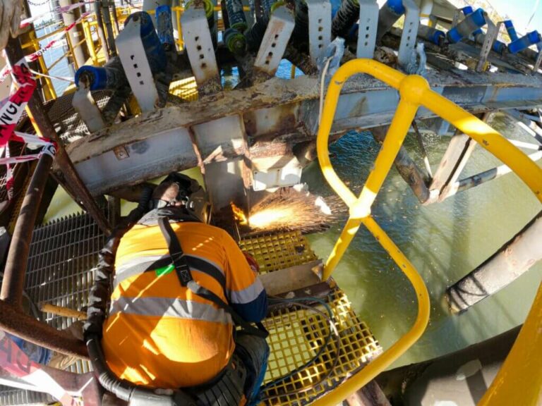 A worker in safety gear and a high-visibility jacket uses a cutting tool to slice through metal beams on an industrial structure above water, demonstrating advanced steel design. The scene is surrounded by yellow safety railings and caution tape, with sparks flying from the fabrication process.