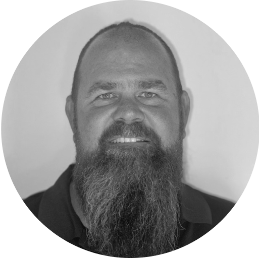 Black and white headshot of a man with a full beard. He is looking straight at the camera and smiling slightly. The background is plain and the image is circularly cropped.