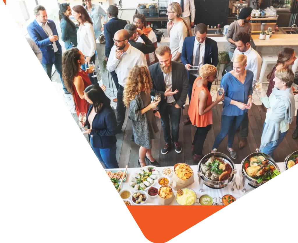 A diverse group of people engaged in conversation at a social event. They are standing and mingling around a buffet table filled with various dishes. Some are holding drinks, while others are deep in discussion. The atmosphere is lively and informal, reflecting the vibrant spirit of their communities.