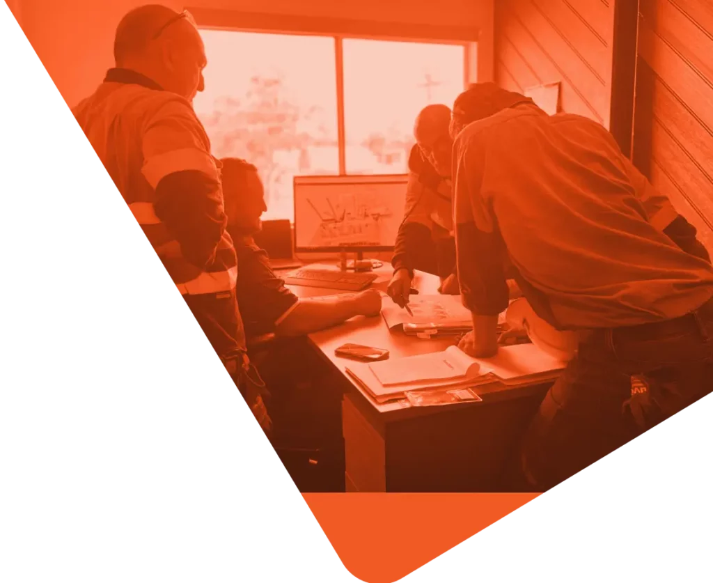 A group of four people in an office are collaborating at a table with documents and a computer. The image, tinted in strong red hues, showcases one person seated while the others stand around, intently reviewing the plans. The atmosphere exudes a sense of focused engagement and vibrant communities coming together.